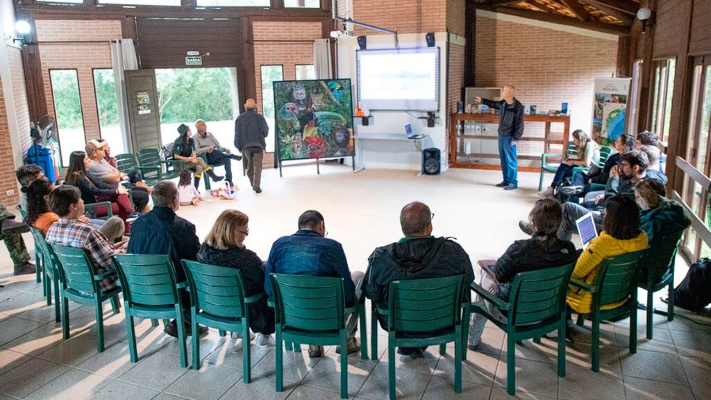 Imagem de pessoas sentadas em cadeiras olhando para um projetor e uma obra de arte com animais e floresta.