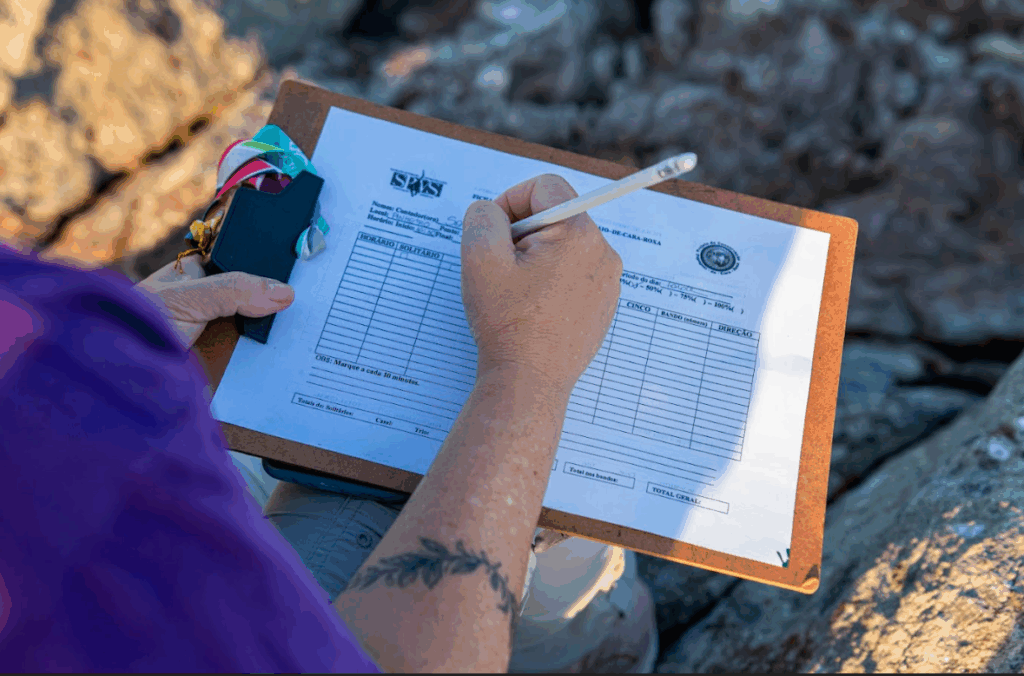 Imagem de uma pessoa preenchendo uma folha com tabela, em cima de uma prancheta.