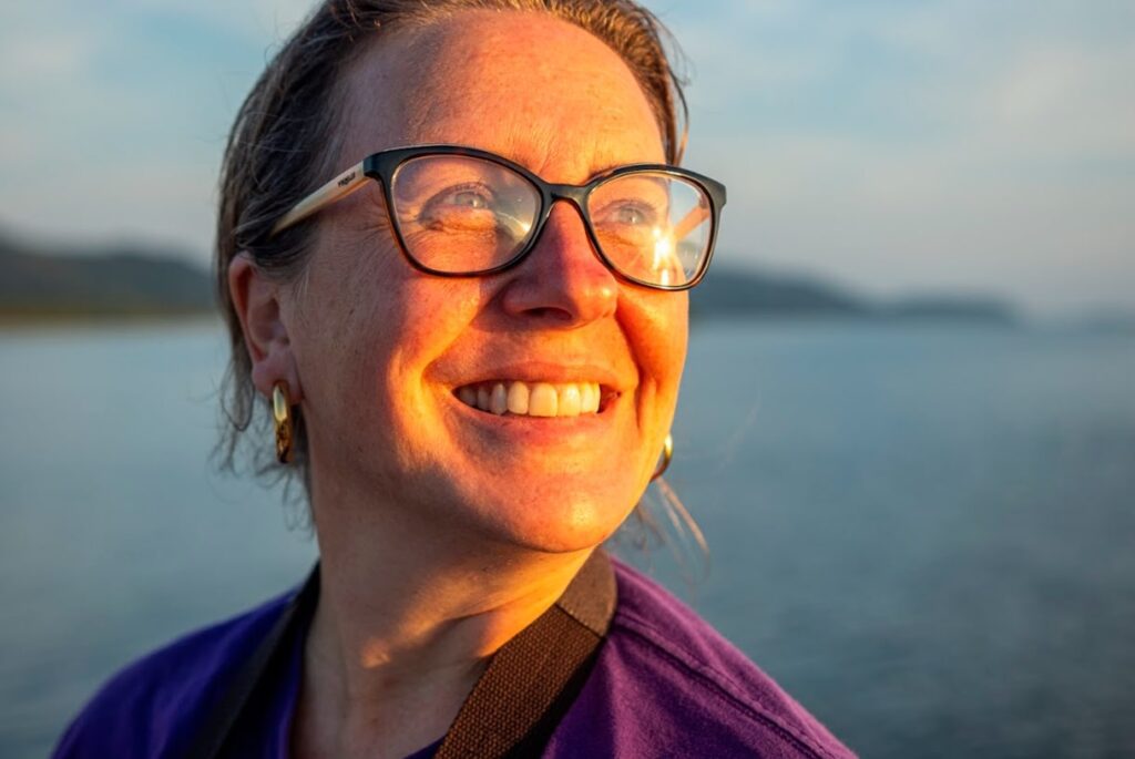 Imagem da Solange Latenek, uma mulher loira, de óculos de grau, sorrindo olhando para o sol.