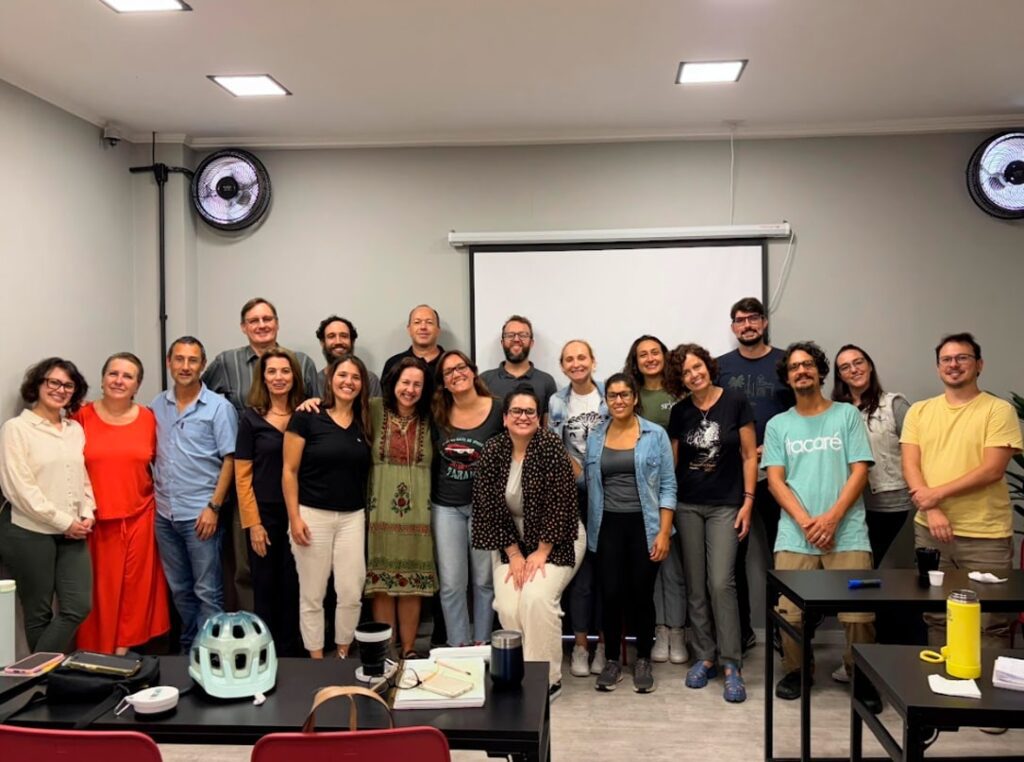 Imagem com 20 integrantes da SPVS e o Ignácio posando para a foto. Ao fundo, uma lousa de projeção e caixas de som na parede.