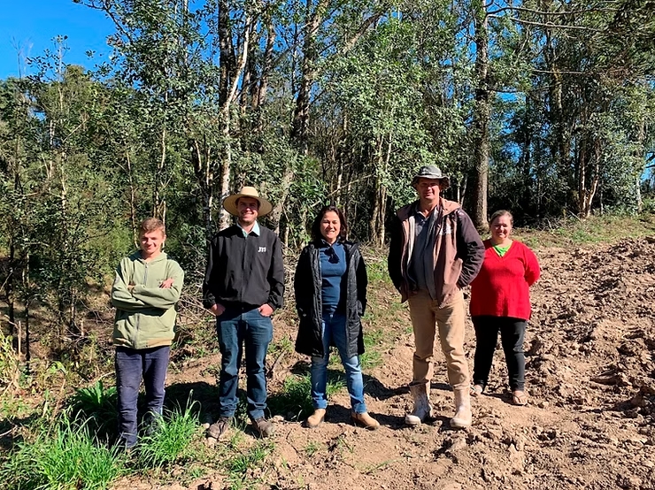 Imagem de cinco pessoas - duas mulheres e três homens - em pé em chão com terra e grama. Ao fundo, árvores e o céu.