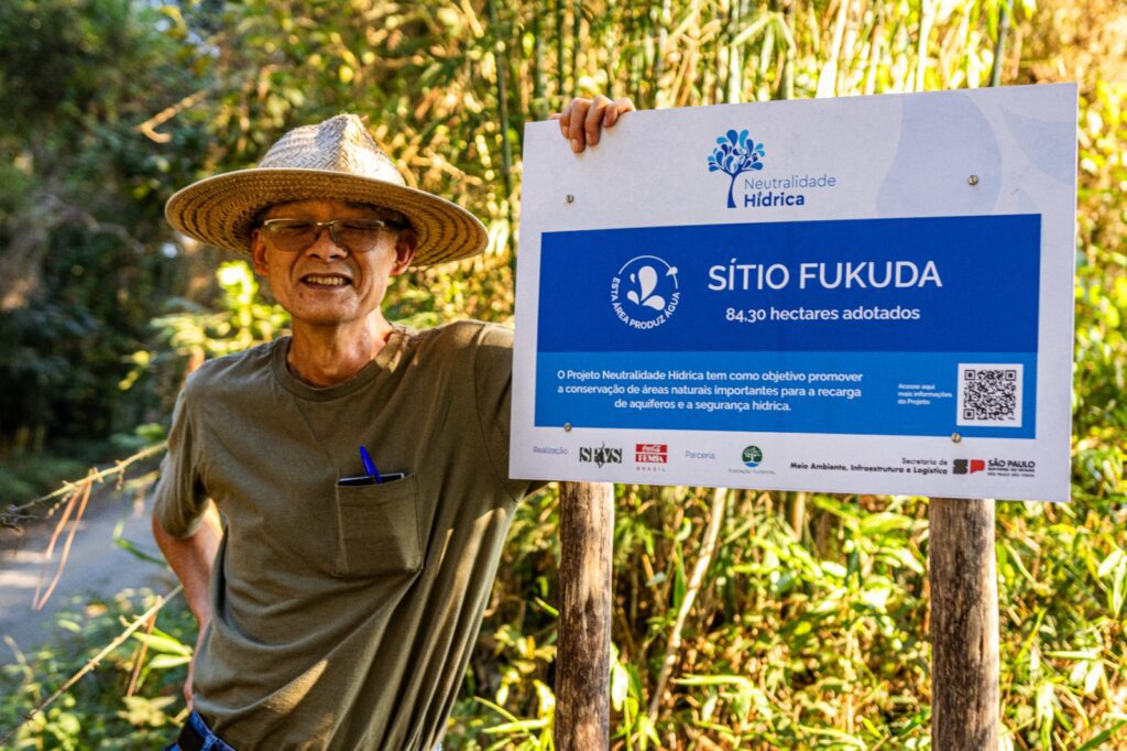Imagem de um homem sorrindo usando um chapéu e apoiado em uma placa com escrito: "Sítio Fukuda".
