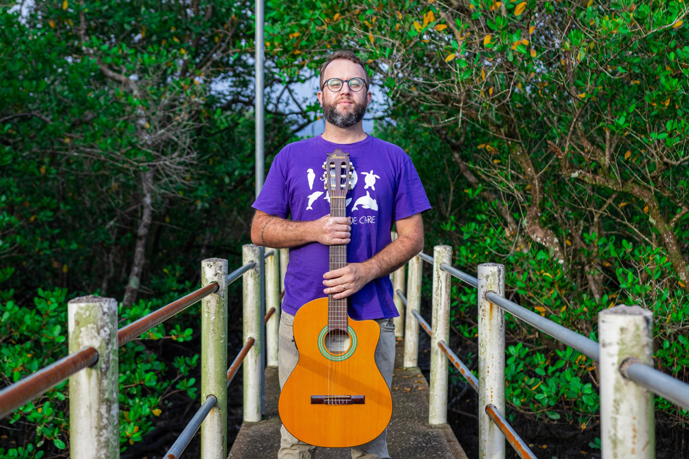Post do Blog da SPVS: Homem de óculos e camiseta roxa segurando um violão em uma passarela ao ar livre, cercada por vegetação.