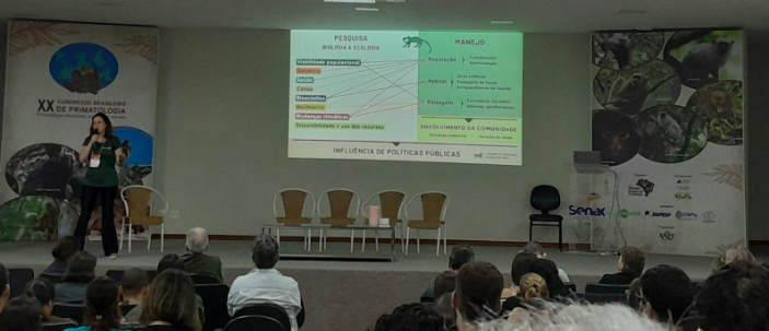 Imagem de uma mulher apresentando uma pesquisa projetada em projeto, para a plateia.