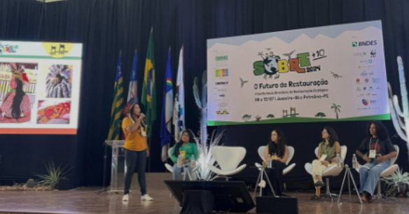 Imagem de uma mulher olhando para pessoas sentadas a frente um banner.