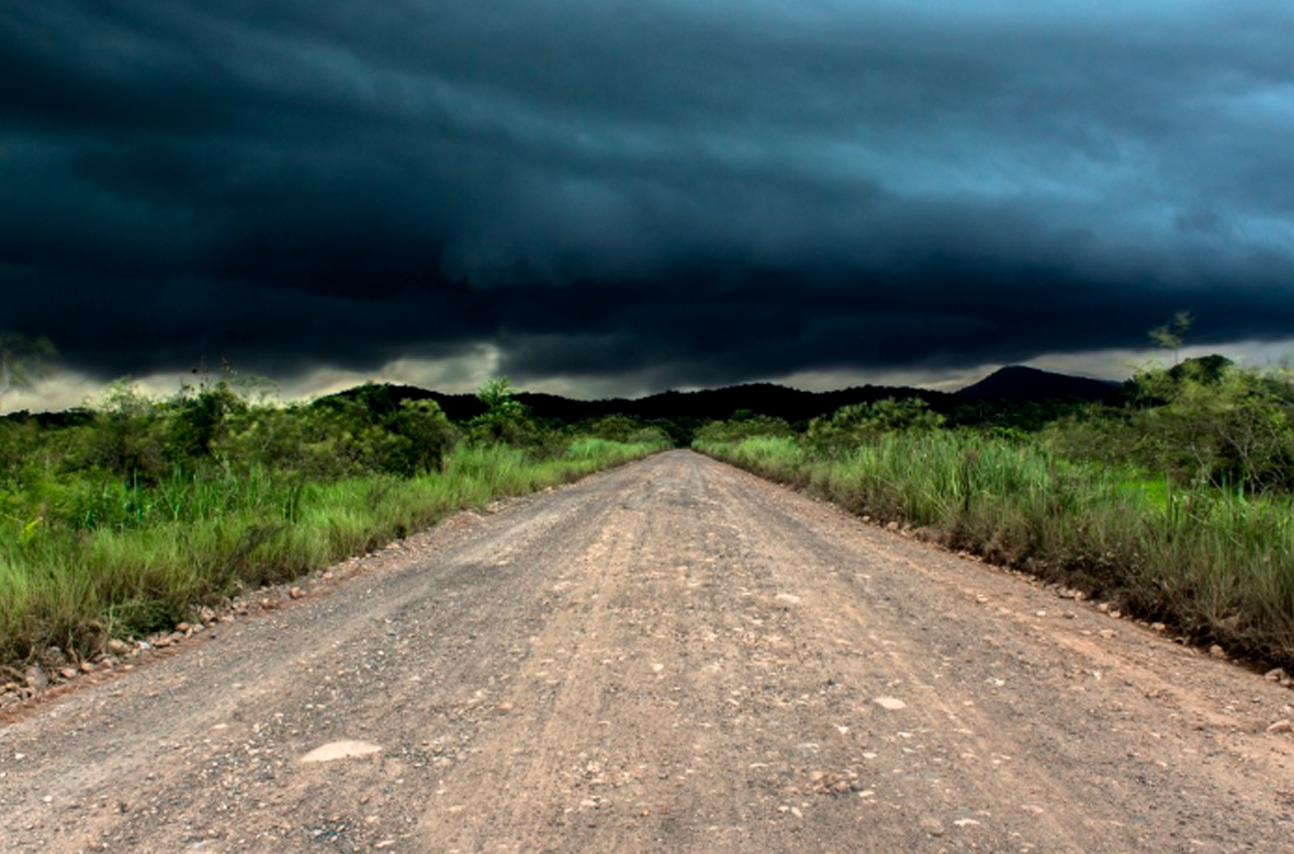 Imagem de uma estrada de terra, com matos no canto. O céu está com muita nuvem e escuro.