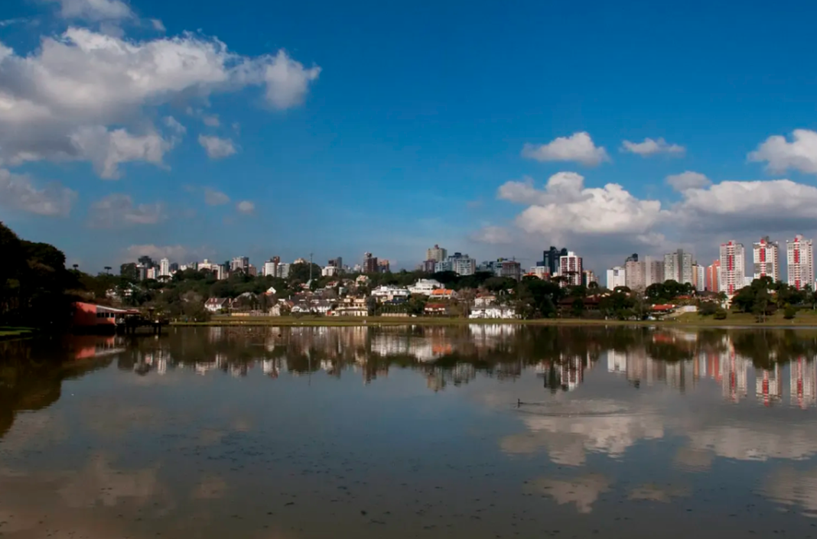 Imagem de um lago refletindo a cidade, árvores e o céu.