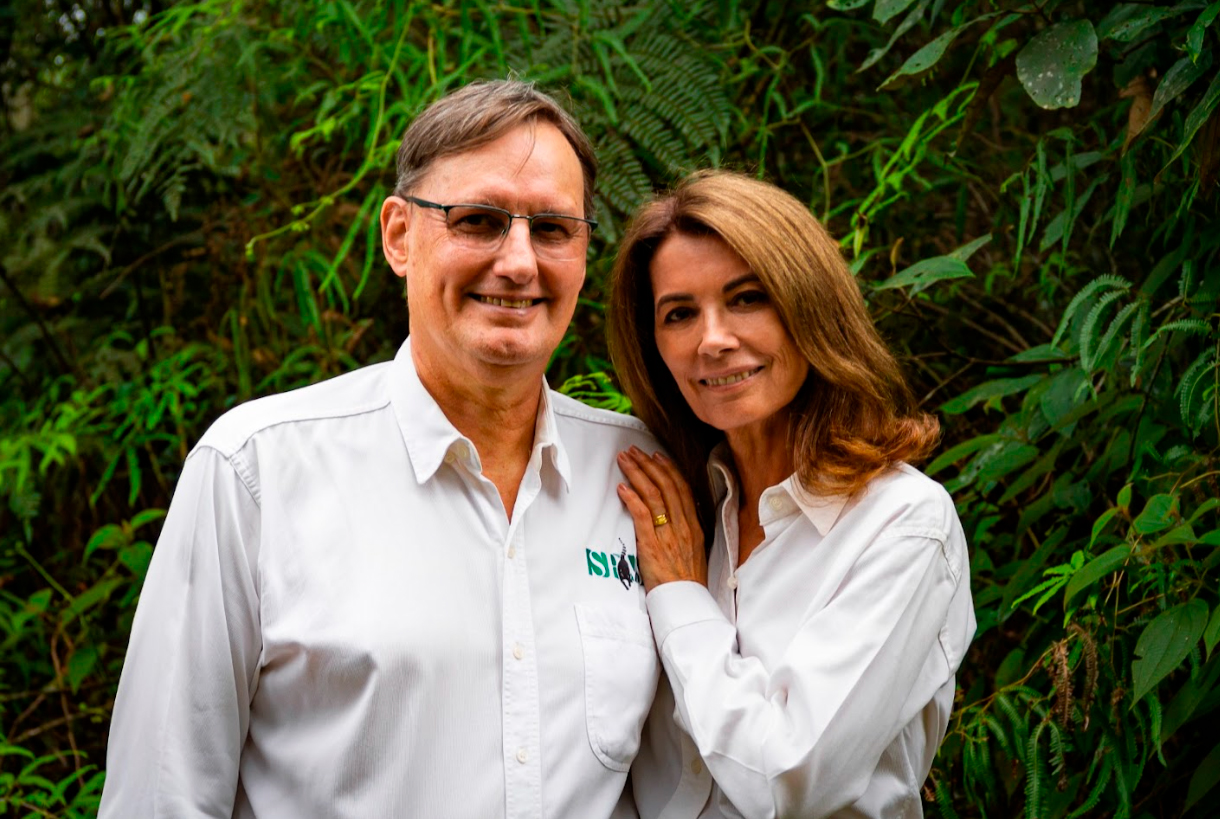 Imagem de Clóvis Borges e Mônica Borges se abraçando e sorrindo para a foto. Ambos são brancos e estão usando camisas sociais brancas. Clóvis usa óculos e tem cabelo liso castanho curto, Mônica tem cabelo liso na altura dos ombros.