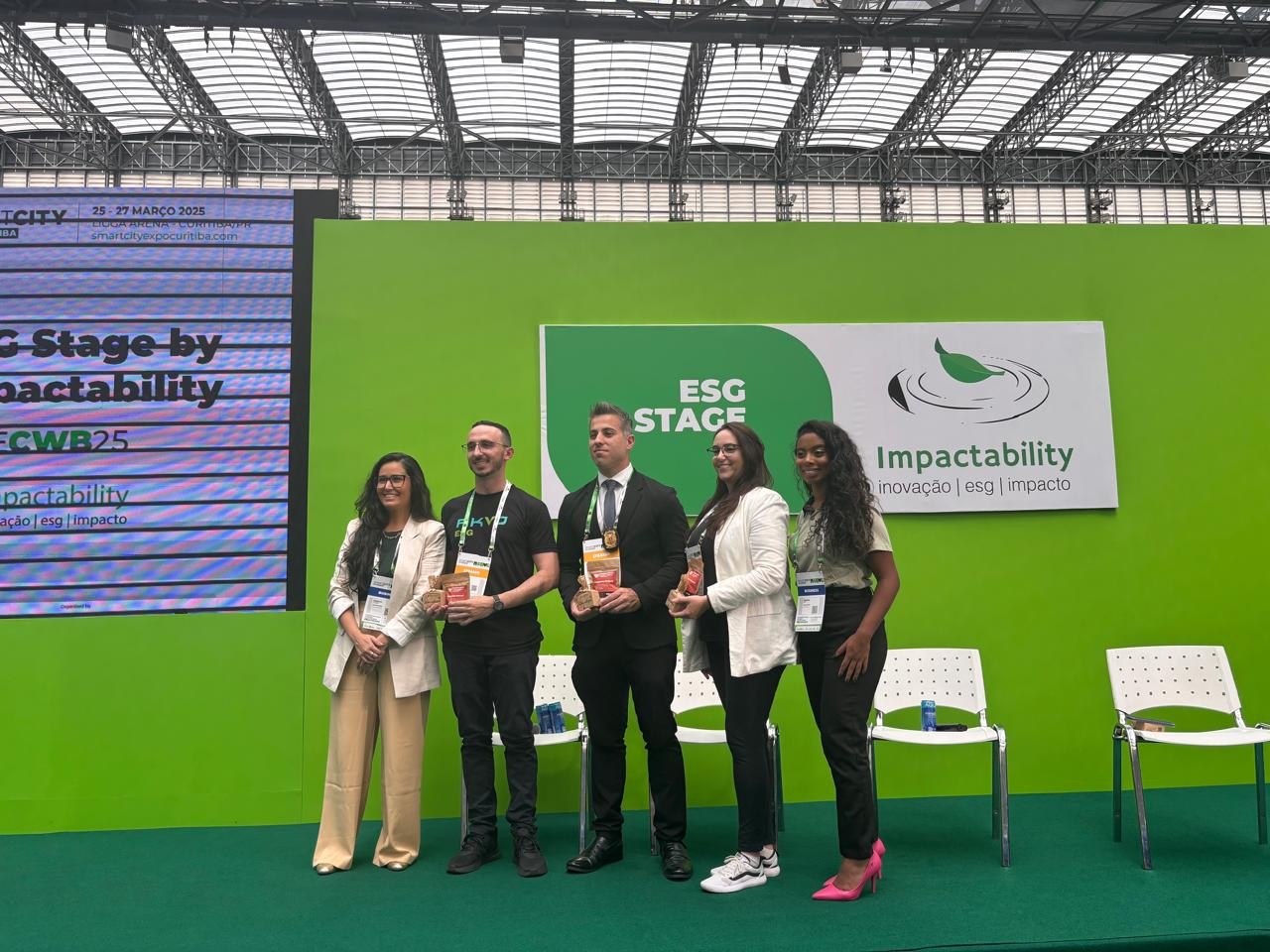 Imagem com cinco pessoas, dois homens e três mulheres, em pé usando roupas formais, crachás. Ao fundo, tem cadeiras e um painel escrito "ESG Stage - Impactbility".